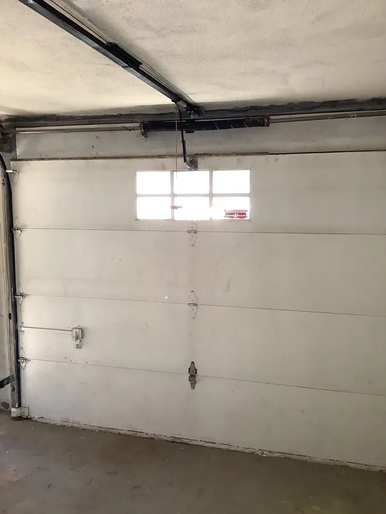 Closed white garage door seen from inside with a single six-light window in the top section and the torsion spring and shaft running across the header above it
