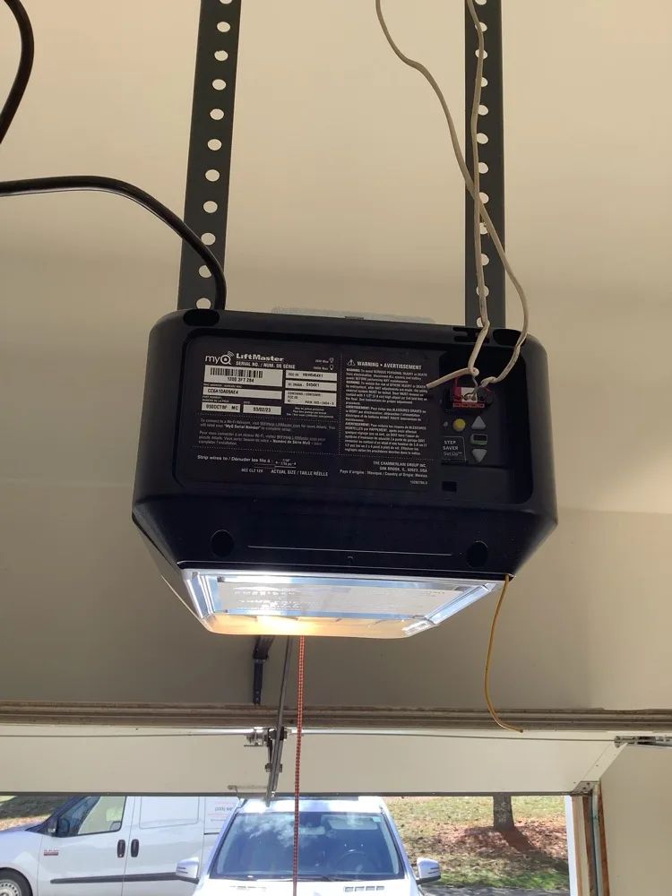 LiftMaster garage door opener head suspended from the ceiling on punched steel hangers, its rating label and lit lens facing down, with the open door and a green lawn beyond