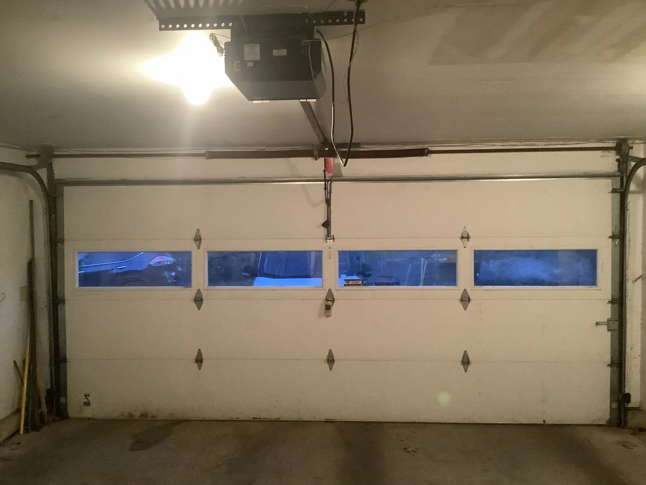 Closed white double garage door seen from inside, a row of four windows across the middle section and a lit chain-drive opener on the ceiling above the header